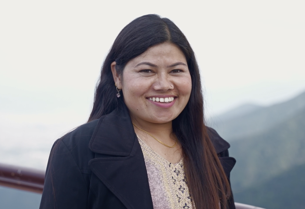 Pampha, a Nepali woman, is smiling and standing outdoors. She has long dark hair and is wearing a black jacket and a Kurt. 
