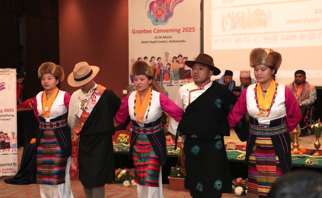 Five Indigenous dancers are dancing inside a conference room. They are wearing traditional dresses and hats.