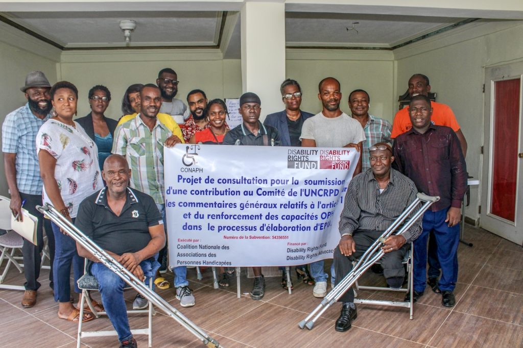 A group of Haitian people are smiling and posing for a photo. They are holding a banner in French.
