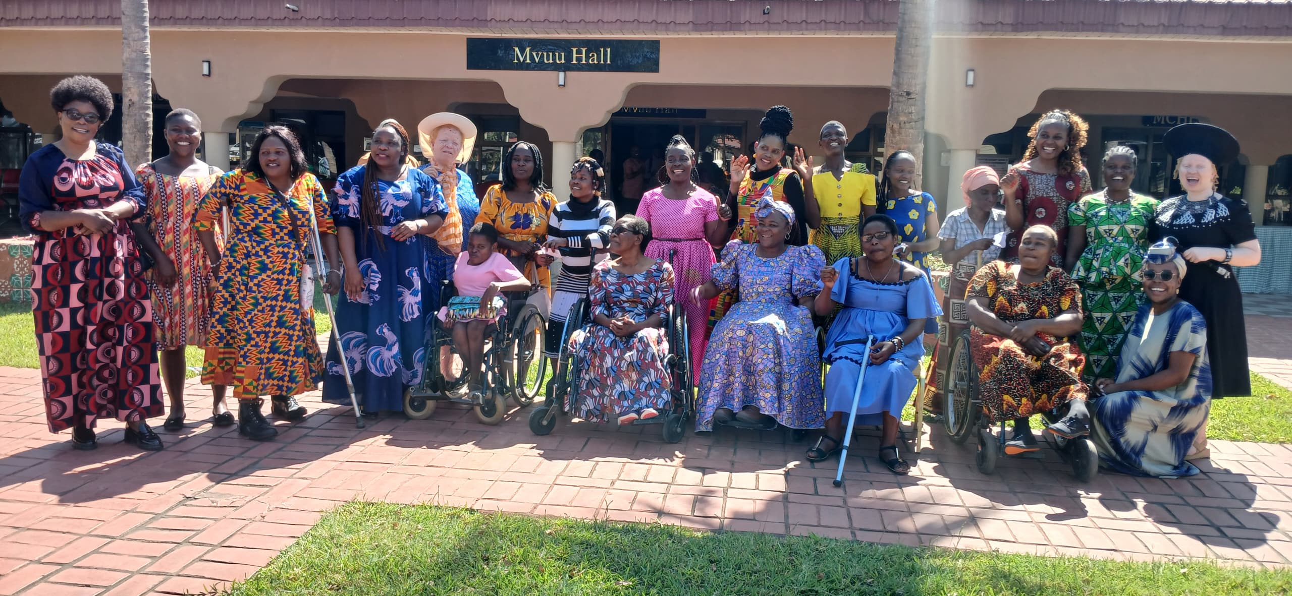 A smiling group of women, some using wheelchairs, gather outside in front of a building.