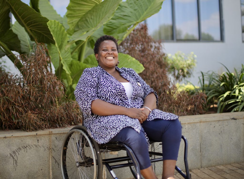 An outdoor photo of a smiling African woman. She's wearing a blue and white printed top and blue pants and is using a wheelchair.