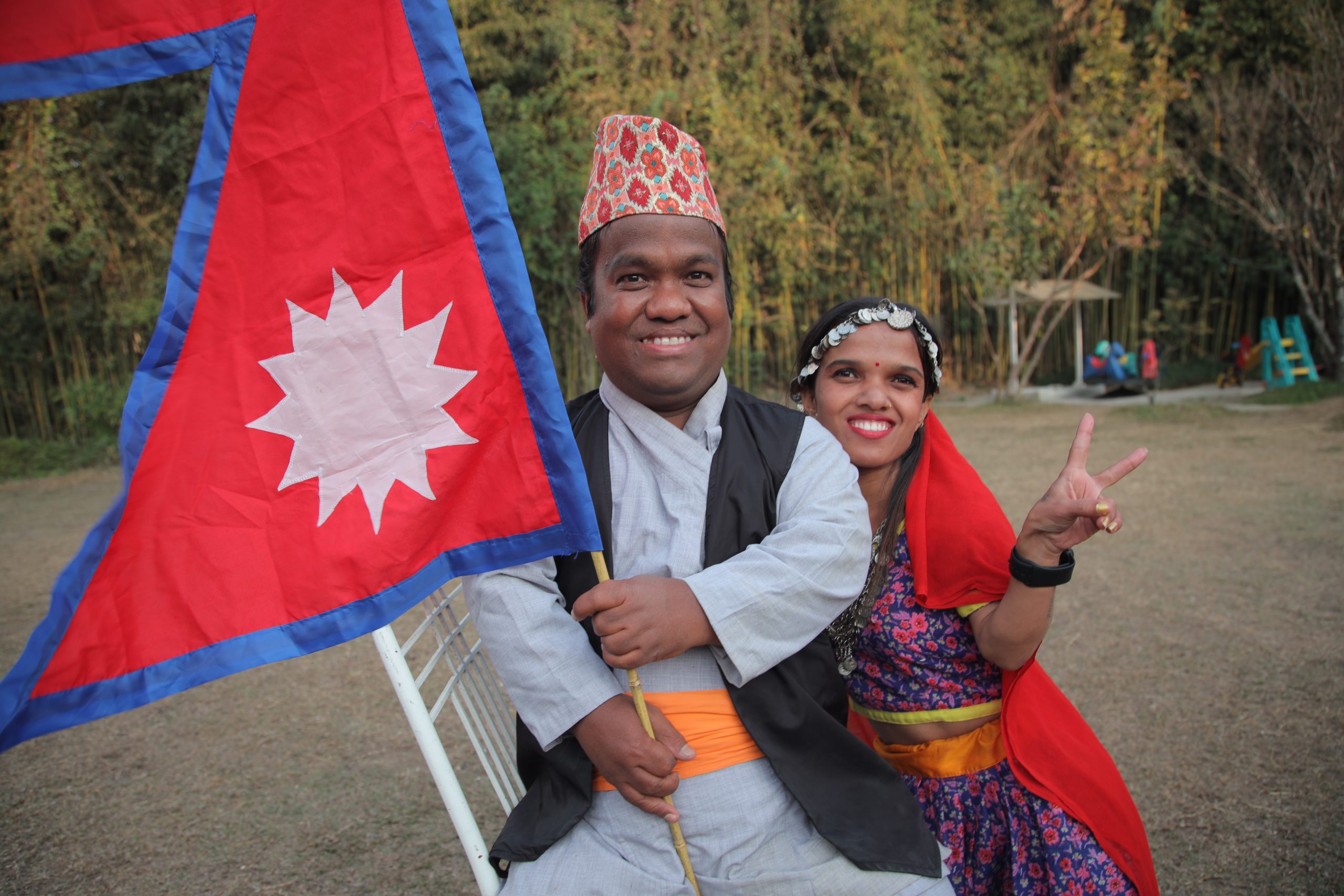 A Nepali man and woman are smiling. The woman is giving a peace sign and the man is holding a flag.