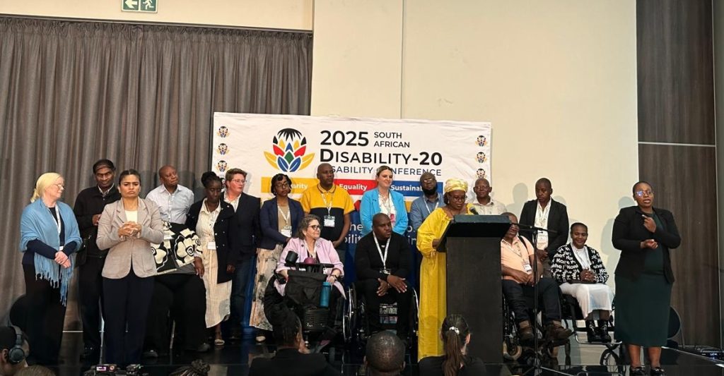 A group of people are gathered on a stage in front of the banner of Disability-20.