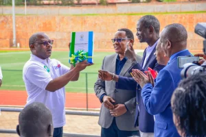 Several men in suits clapping and smiling while a man in a white t-shirt holds up the Rwandan Sign Language Dictionary which is wrapped in a colourful bow